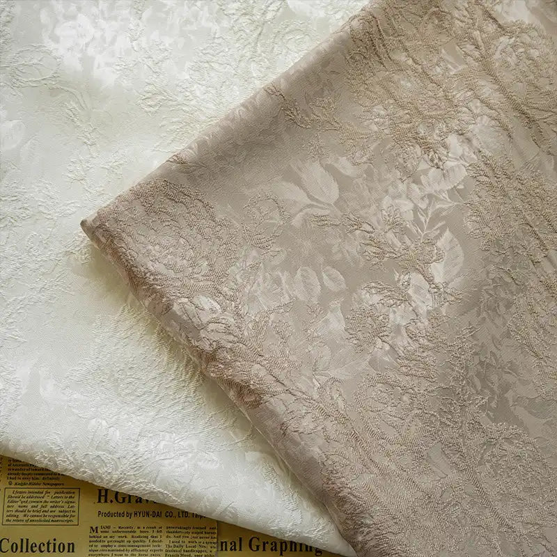 Two pieces of Longan Craft’s Embossed Peony Brocade Fabric—one ivory, one light beige—featuring a subtle Chinese-style floral pattern, are side by side on newspaper. This elegant fabric adds refinement to dresses or any DIY project.