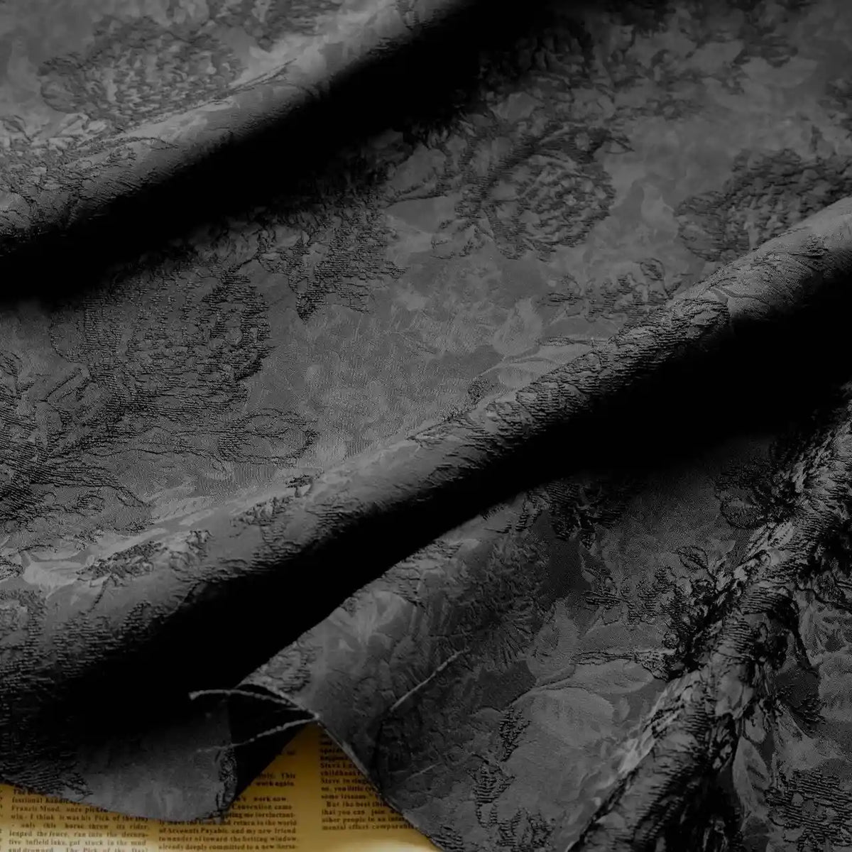 A close-up of Longan Craft's Embossed Peony Brocade Fabric By The Yard, a black Chinese style floral brocade for dresses, draped in soft folds with part of a yellowed newspaper peeking underneath at the edge.
