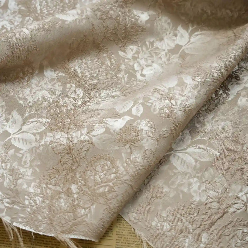 A close-up of Longan Craft's Embossed Peony Brocade Fabric By The Yard, a Chinese-style floral brocade for dresses, in light beige with an intricate pattern, softly draped with frayed edges and printed paper partially beneath.