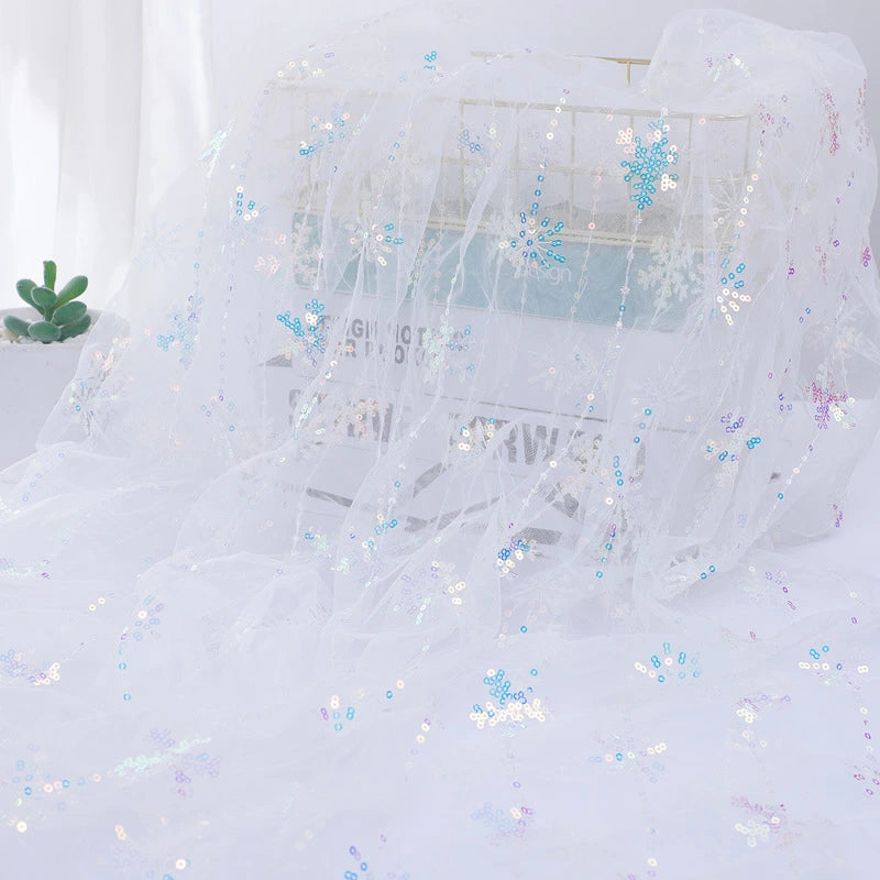 Longan Craft's Embroidered Sequin Tulle Fabric, featuring sparkling snowflake embroidery, is draped over a wire basket. A small green succulent sits nearby, softly illuminated from the left. Perfect for princess dress material.