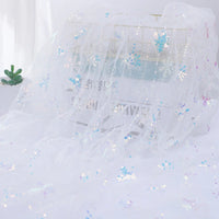 Longan Craft's Embroidered Sequin Tulle Fabric, featuring sparkling snowflake embroidery, is draped over a wire basket. A small green succulent sits nearby, softly illuminated from the left. Perfect for princess dress material.