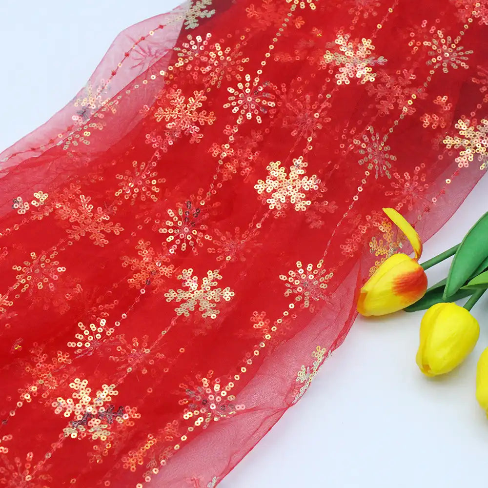 A close-up of Longan Craft's Embroidered Sequin Tulle Fabric by the yard, featuring sheer red tulle with sparkling gold snowflake embroidery, beside three yellow tulips with red tips on a white surface.