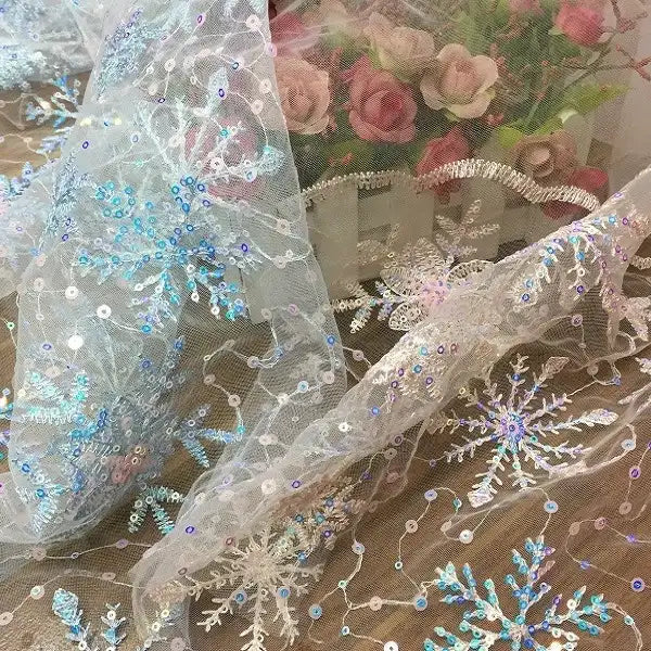 Longan Craft’s Embroidered Sequin Snowflake Tulle Fabric is draped in front of a white basket filled with pink and cream artificial roses, showcasing its blue and silver snowflake embroidery, perfect for DIY wedding dresses or children’s apparel.