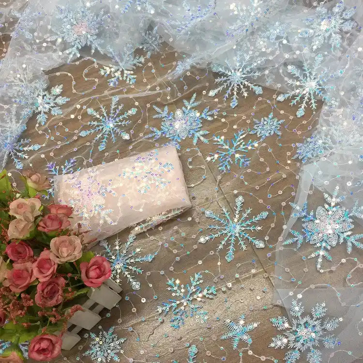 Longan Craft’s Embroidered Sequin Tulle Fabric, featuring sparkling blue and silver snowflakes, is displayed with a light pink sequin clutch and a small bouquet of pink roses. Perfect for DIY wedding dresses and children’s apparel.