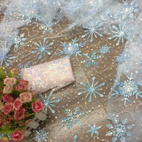 Longan Craft’s Embroidered Sequin Tulle Fabric, featuring sparkling blue and silver snowflakes, is displayed with a light pink sequin clutch and a small bouquet of pink roses. Perfect for DIY wedding dresses and children’s apparel.