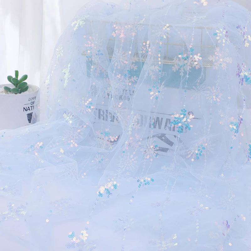 Longan Craft's Embroidered Sequin Tulle Fabric By The Yard features sparkling snowflake embroidery, shown draped over a book and a succulent, with soft natural light highlighting its princess dress-worthy shimmer.