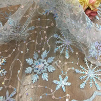 A close-up of Longan Craft's Embroidered Sequin Tulle Fabric by the yard, featuring snowflake embroidery and sparkling sequins—perfect for DIY wedding dresses and children’s apparel—with soft pink and yellow flowers in the background.