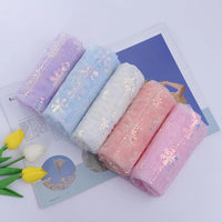 Five rolls of Longan Craft Embroidered Sequin Tulle Fabric (purple, blue, white, peach, pink) with sparkling snowflake embroidery are displayed on an open magazine beside yellow and white tulips. Ideal for princess dresses.