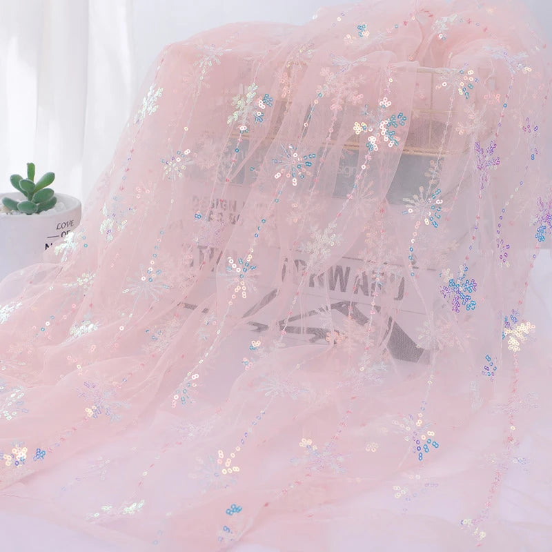 Longan Craft’s Embroidered Sequin Tulle Fabric By The Yard, featuring sparkling snowflake designs, is draped over a clear box. A small green potted succulent sits nearby on a white surface.