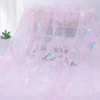 Longan Craft’s Embroidered Sequin Tulle Fabric By The Yard features sparkling snowflake embroidery in sheer pink, draped over a wire rack with a newspaper and potted succulent in the background—perfect for princess dress material.