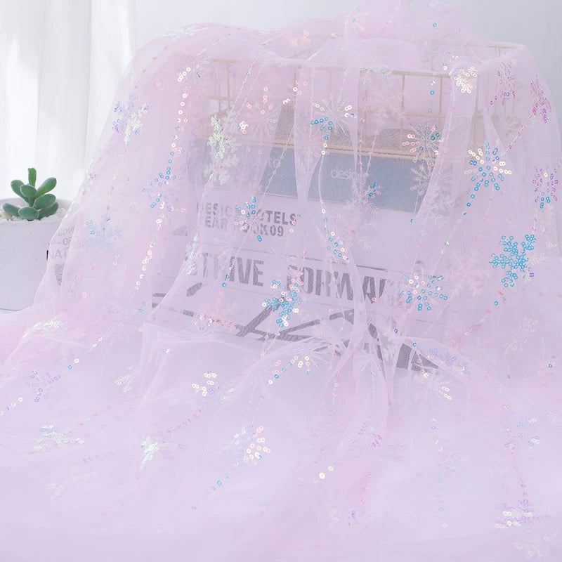 Longan Craft’s Embroidered Sequin Tulle Fabric By The Yard features sparkling snowflake embroidery in sheer pink, draped over a wire rack with a newspaper and potted succulent in the background—perfect for princess dress material.