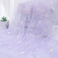 Longan Craft’s Embroidered Sequin Tulle Fabric By The Yard features sparkling snowflake embroidery and iridescent sequins in light purple, draped over a surface with a potted succulent on the left and blurred text visible through the fabric.