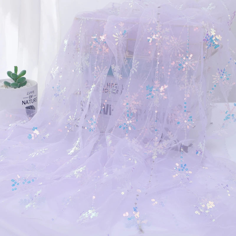 Longan Craft’s Embroidered Sequin Tulle Fabric By The Yard features sparkling snowflake embroidery and iridescent sequins in light purple, draped over a surface with a potted succulent on the left and blurred text visible through the fabric.