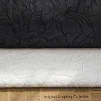 Two rolls of Longon Craft Floral Embossed Jacquard Brocade Fabric—one black, one white—lie side by side on newspaper, showcasing a raised 3D solid flower pattern in polyester for party costumes and tailored suits.