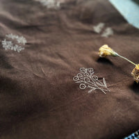 A piece of Longan Craft’s Floral Embroidered Cotton Corduroy Fabric By The Yard in brown lies flat, showing off its soft pinwale corduroy texture and white stitched floral designs with two dried flowers on the side.