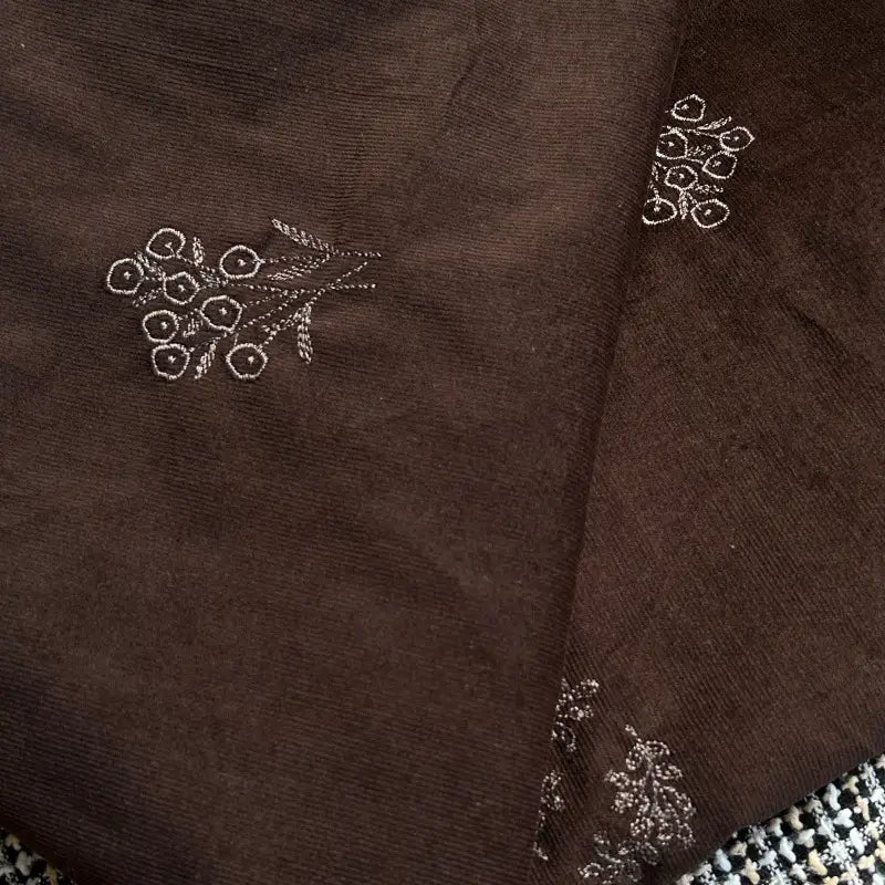 Longan Craft Floral Embroidered Cotton Corduroy Fabric by the Yard in brown features fine pinwale texture and small white floral designs. This soft autumn textile is slightly folded, displaying part of its patterned surface in the corner.