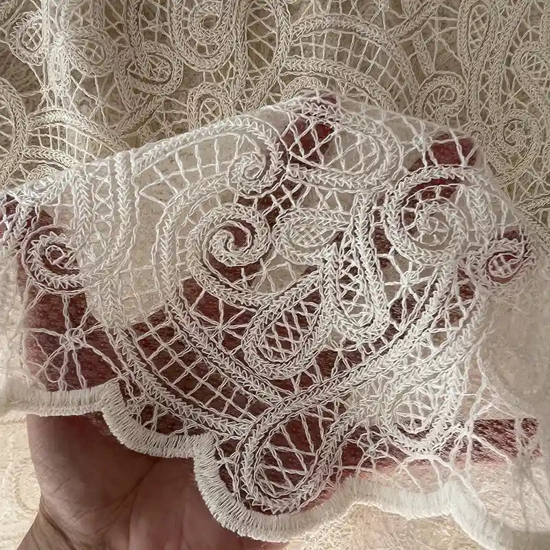 A hand displays Longan craft’s Floral Embroidered Lace Fabric By The Yard—cotton thread fabric with intricate floral embroidery, ideal for wedding dresses. Light filters through the delicate off-white material.