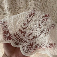 A hand displays Longan craft’s Floral Embroidered Lace Fabric By The Yard—cotton thread fabric with intricate floral embroidery, ideal for wedding dresses. Light filters through the delicate off-white material.