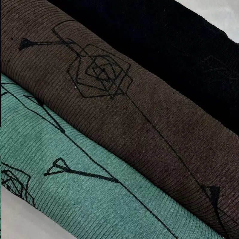 Three rolls of Longan Craft Floral Embroidered Polyester Corduroy Fabric by the yard in black, brown, and turquoise are stacked diagonally, each showing fine wale material with black geometric flower and arrow designs.
