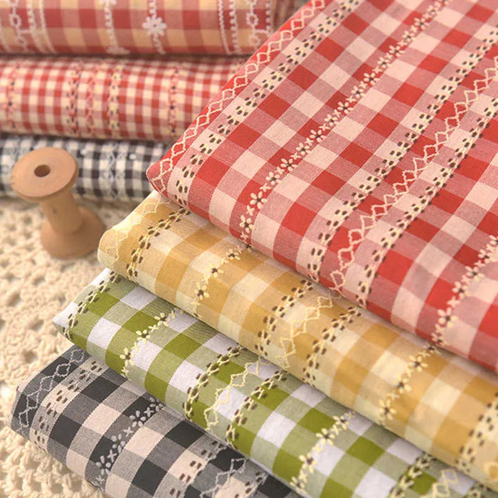 A stack of Longan Craft Floral Gingham Polyester Poplin Fabric By The Yard in red, pink, yellow, green, and black rests on a crocheted surface beside a wooden spool—perfect for women's clothing and craft projects.