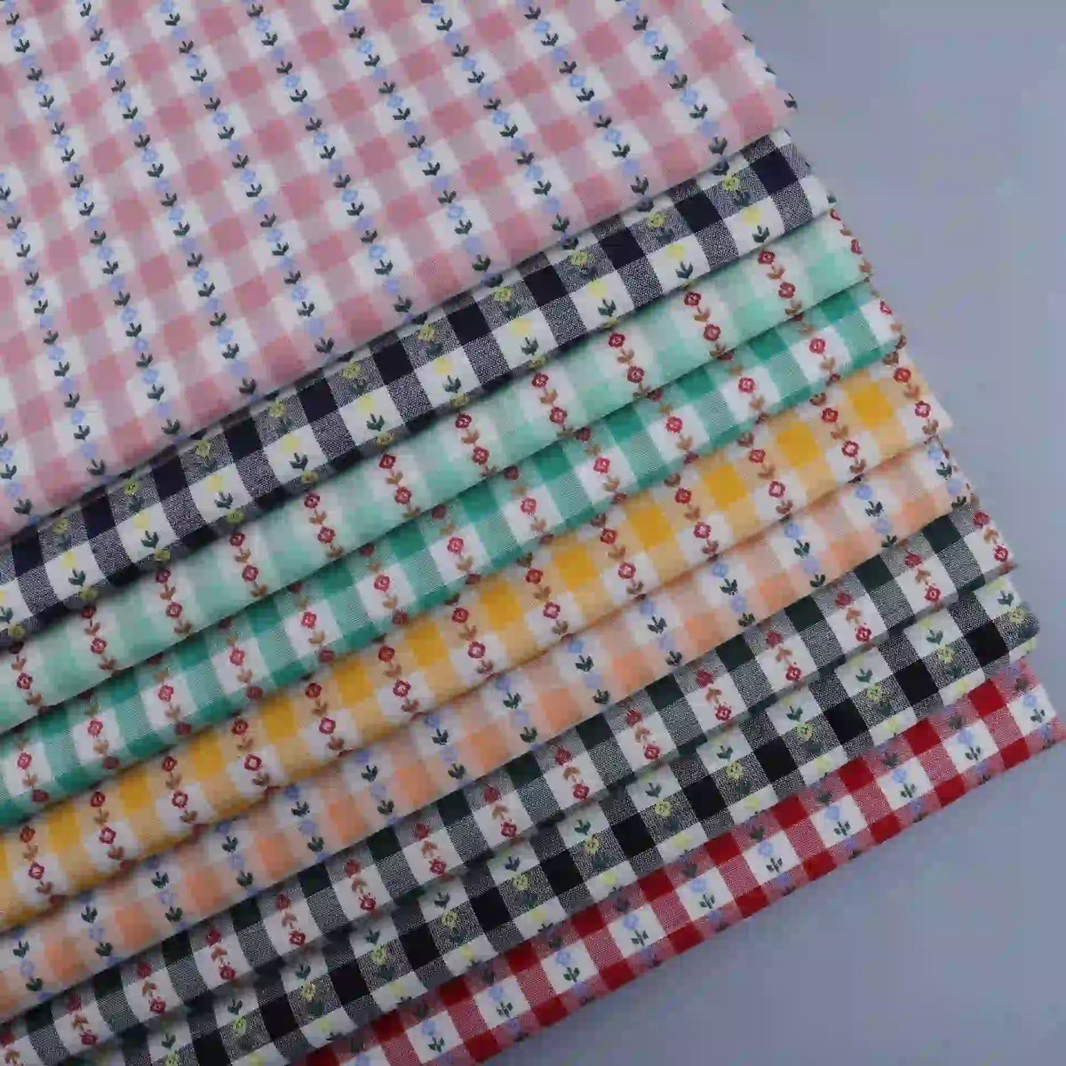 A stack of Floral Gingham Poplin fabrics by Longan Craft, featuring colorful cotton checkered jacquard poplin with charming small floral patterns, is displayed on a light gray surface. These delightful fabrics by the yard are perfect for adding a playful touch to any garment-making or sewing project.