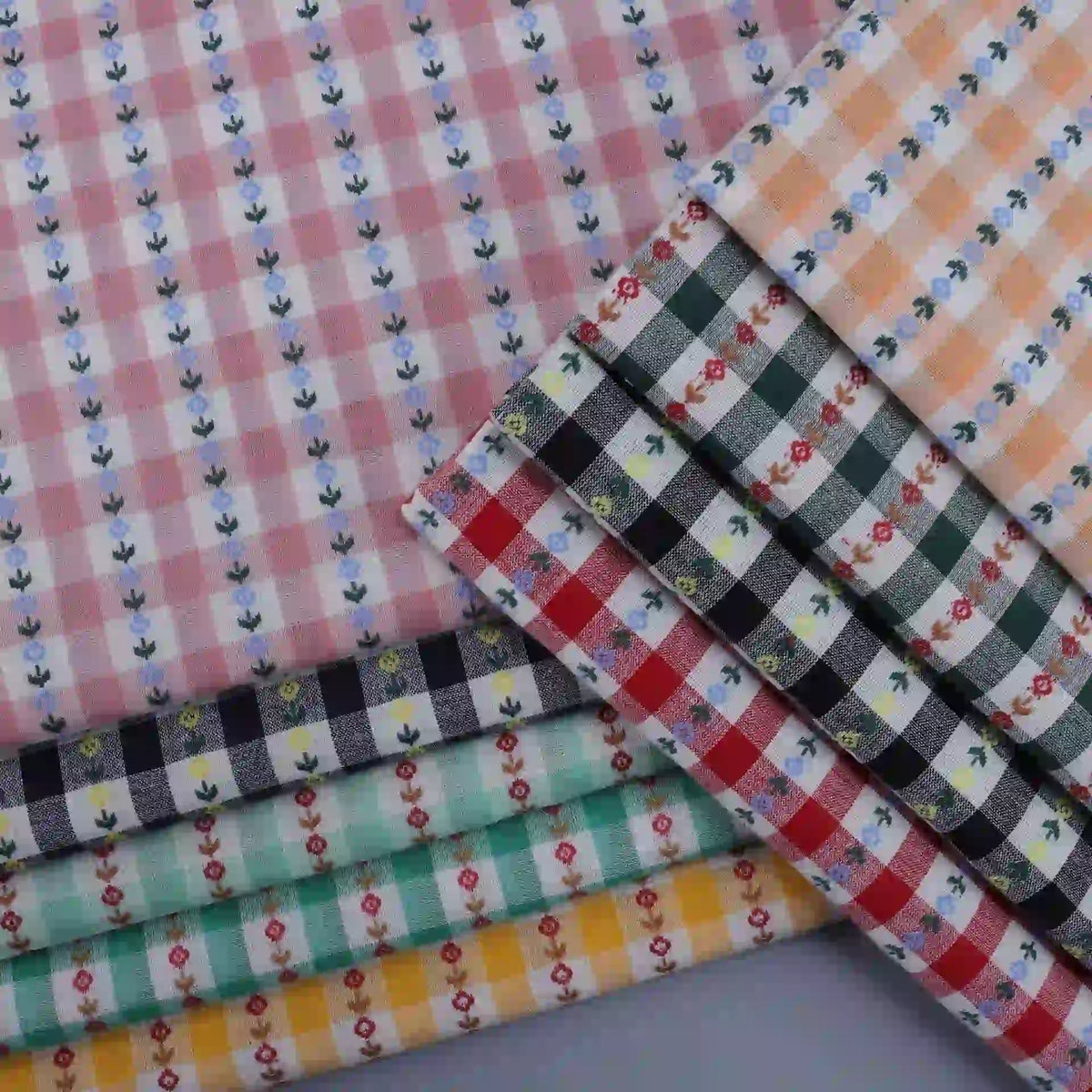 A neatly arranged stack of Longan Craft’s Floral Gingham Poplin Fabric By The Yard showcases a variety of colorful cotton checkered jacquard poplin materials, each featuring delicate small floral designs—making them perfect for garment making.