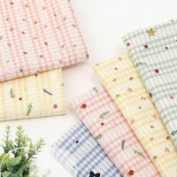 Six pieces of Longan Craft Floral Gingham Quilting Fabric By The Yard, featuring colorful botanical polyester patterns, are displayed on a white surface beside a green plant—ideal for quilting or casual wear projects.