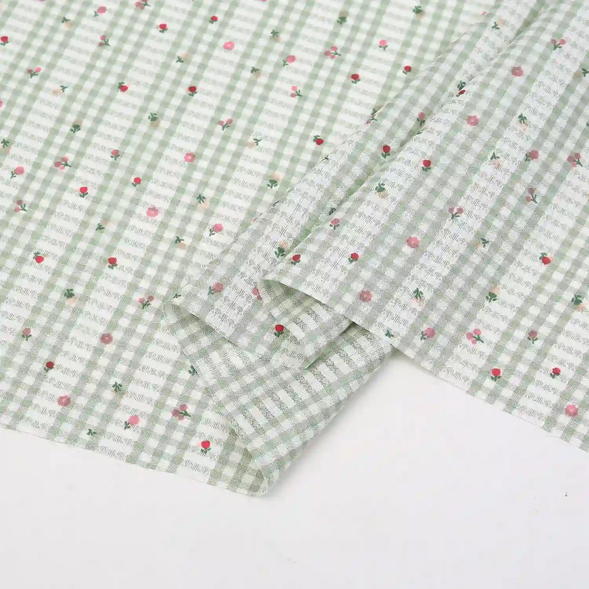 The Longan Craft Floral Gingham Quilting Fabric by the Yard is a comfy polyester fabric in light green and white with a botanical pattern of small pink and red flowers, perfect for quilting or casual wear. One corner is neatly folded.