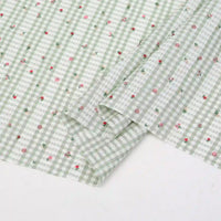 The Longan Craft Floral Gingham Quilting Fabric by the Yard is a comfy polyester fabric in light green and white with a botanical pattern of small pink and red flowers, perfect for quilting or casual wear. One corner is neatly folded.