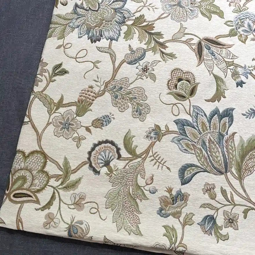 A folded piece of Longan Craft Floral Jacquard Chenille Fabric By The Yard, a heavyweight poly upholstery fabric with an ornate floral pattern in muted green, blue, and beige displayed on a dark grey background—perfect for home decor.