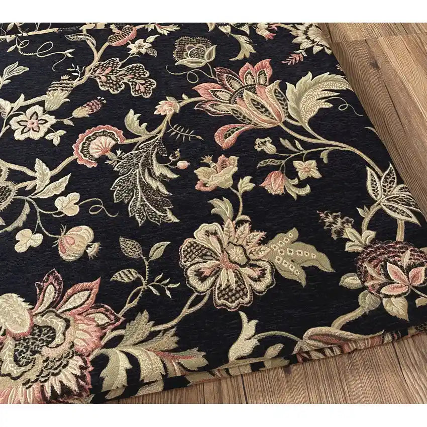 A corner of Longan Craft’s Floral Jacquard Chenille Fabric By The Yard, a heavyweight poly upholstery fabric with an intricate pink, beige, and green pattern on dark background, shown on a wooden floor.