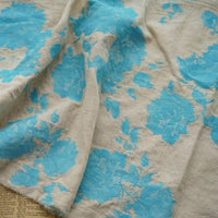 Longan Craft’s Floral Jacquard Cotton Linen Fabric By The Yard features multicolor botanical flower motifs in beige and blue, loosely draped to show its texture. A hint of newspaper peeks from under the fabric—ideal DIY material for dressmaking.