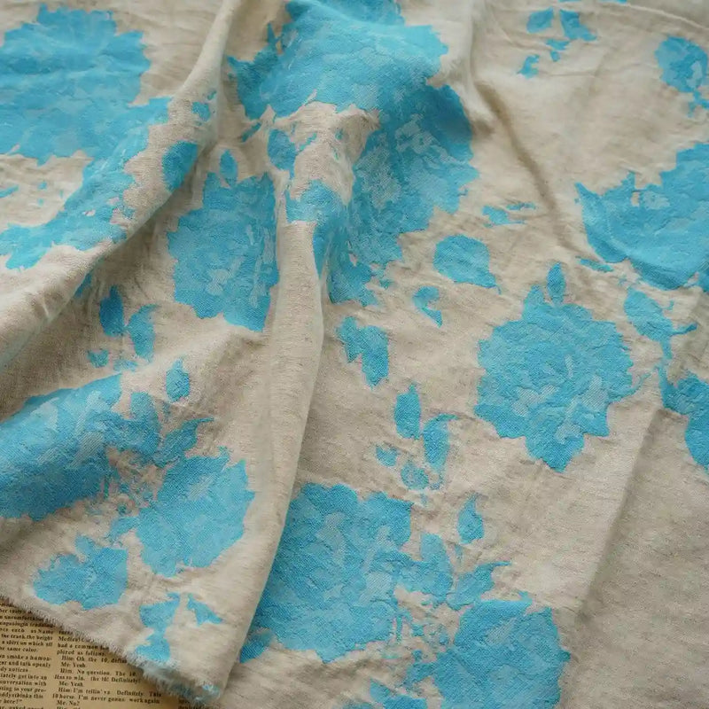 Longan Craft’s Floral Jacquard Cotton Linen Fabric By The Yard features multicolor botanical flower motifs in beige and blue, loosely draped to show its texture. A hint of newspaper peeks from under the fabric—ideal DIY material for dressmaking.