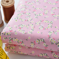 Two folded pieces of Floral Lawn Fabric by the yard from Longan Craft, featuring a pink base with white and yellow floral patterns. Lightweight, breathable cotton lawn fabric. Brown thread spools and a yellow measuring tape are beside it.
