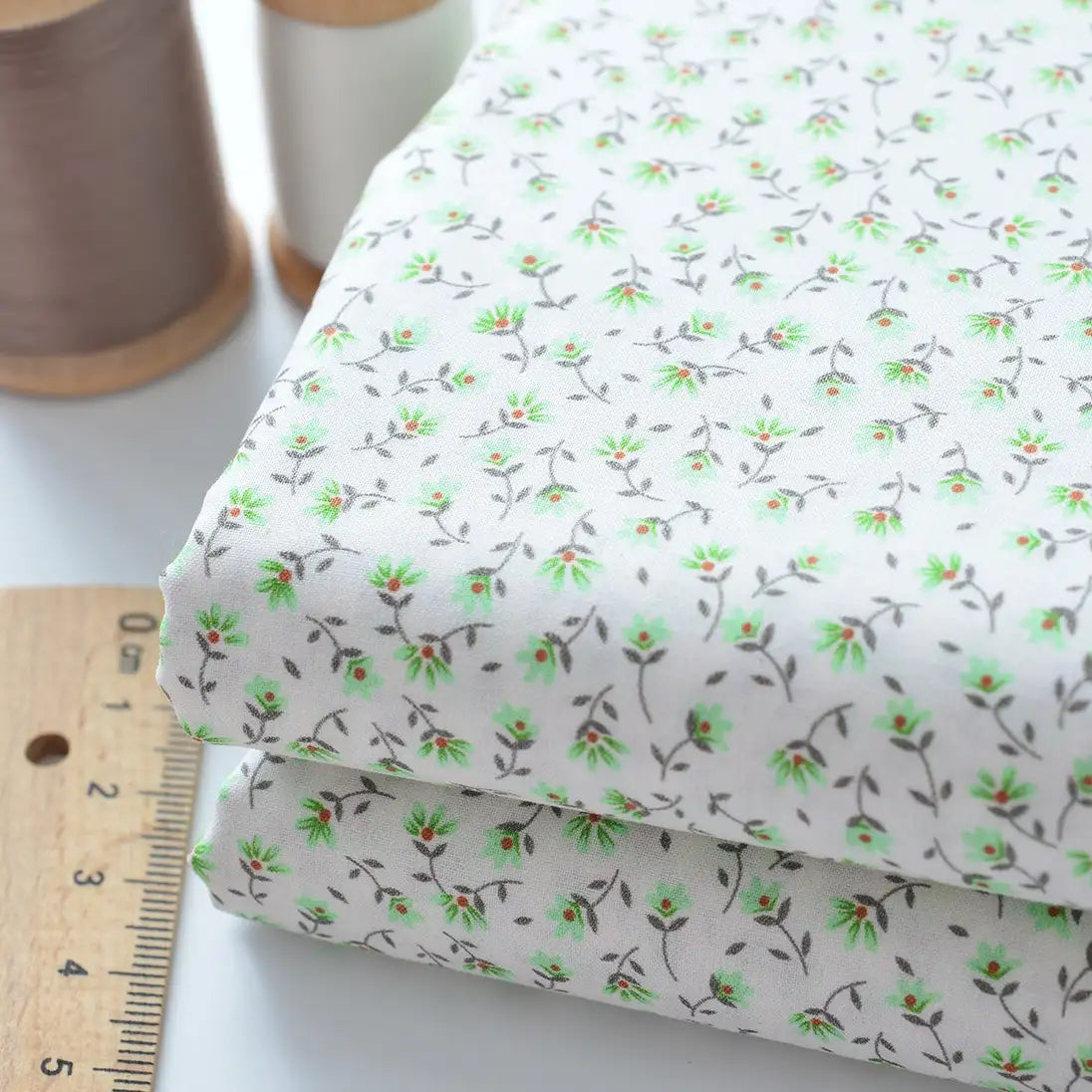 Two pieces of Longan Craft Floral Lawn Fabric By The Yard, lightweight cotton with a green and brown floral pattern, are shown next to a wooden ruler and two spools of thread. Ideal for breathable dress or lining projects.