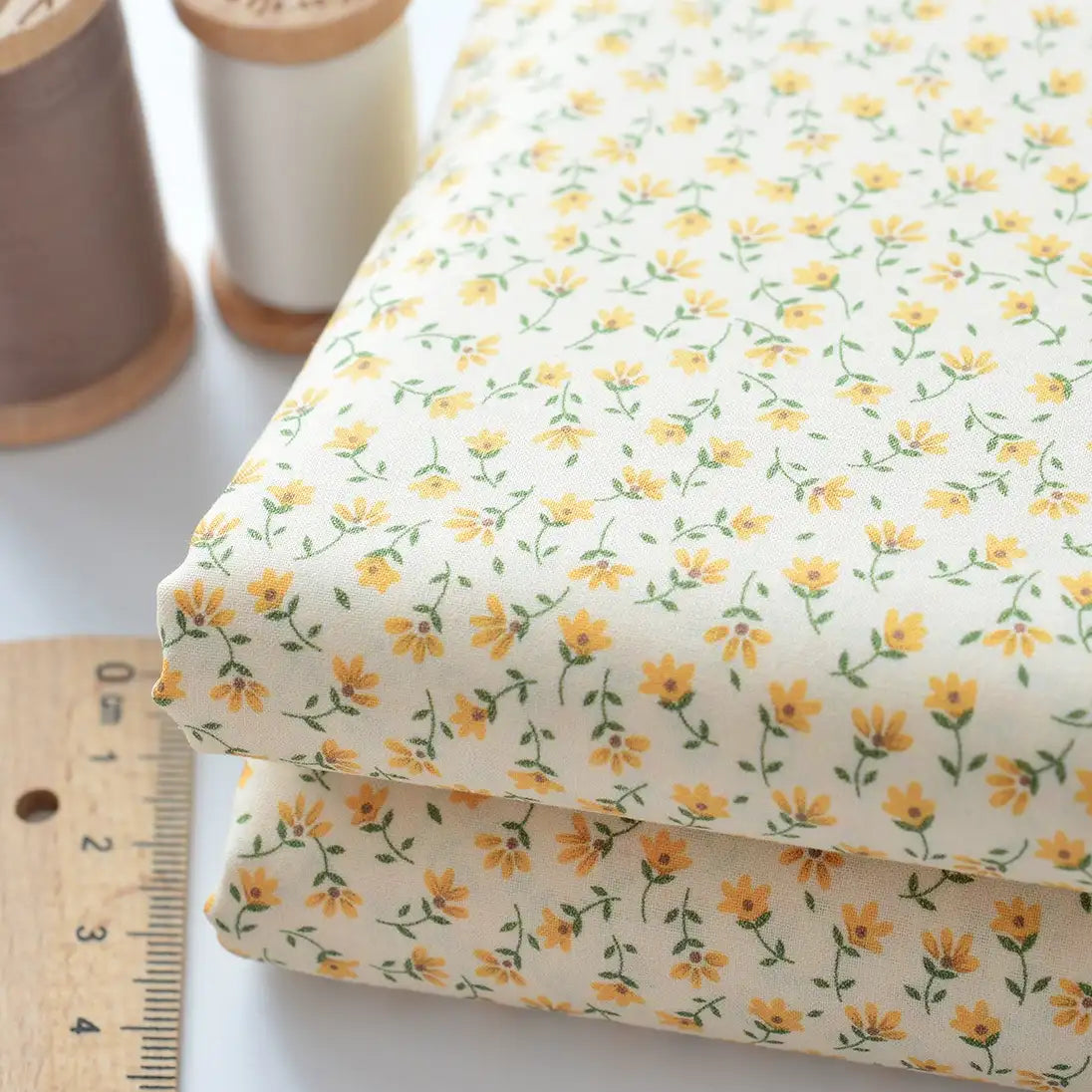 Two folded pieces of Longan Craft Floral Lawn Fabric by the yard—lightweight cotton lawn with a small yellow floral pattern—are shown next to a ruler and two spools of thread, ideal for lining or light sewing projects.