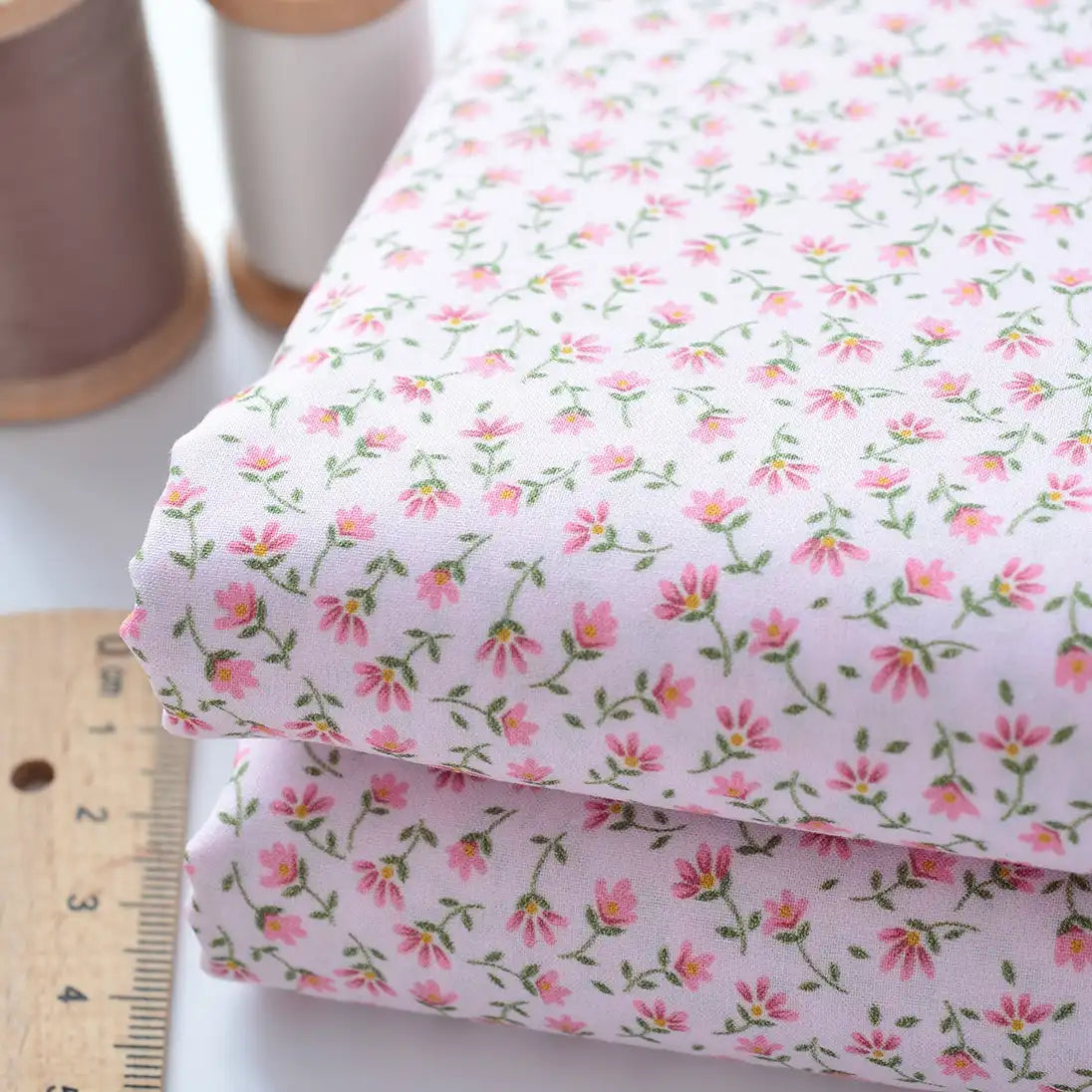 Longan Craft’s Floral Lawn Fabric By The Yard: two folded pieces of lightweight cotton lawn in a delicate pink floral print, with tan and white thread spools and a wooden ruler. Perfect breathable fabric for dresses or linings.