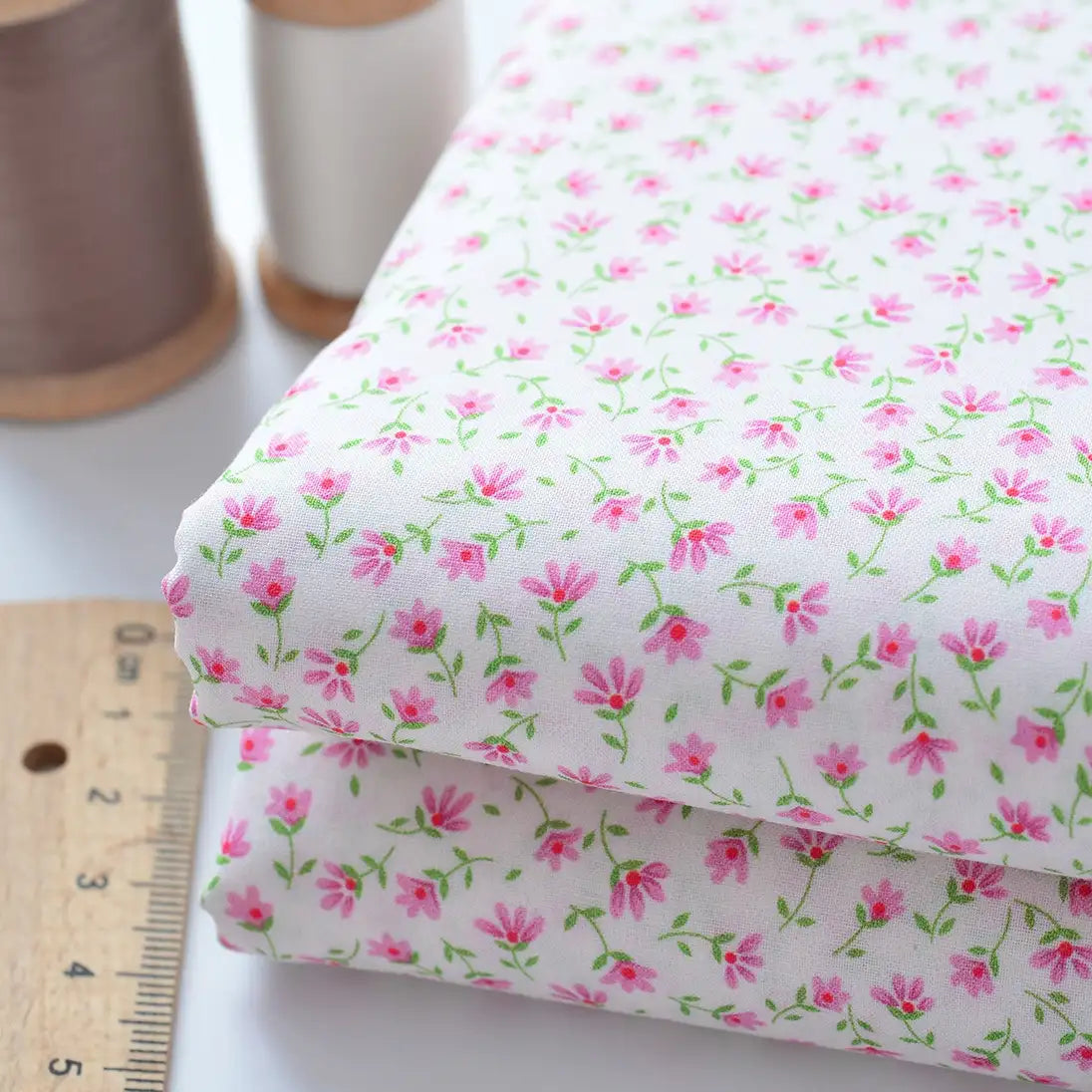 Two pieces of Floral Lawn Fabric By The Yard by Longan Craft in a pink floral pattern are displayed beside a ruler, with spools of thread in the background. This lightweight cotton lawn fabric is perfect for lining and sewing projects.