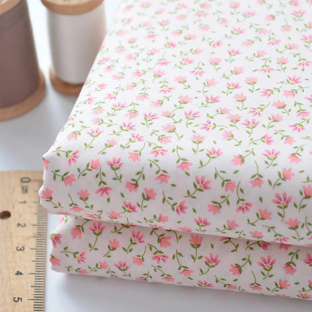 Two pieces of Longan Craft’s Floral Lawn Fabric By The Yard (lightweight cotton with a pink floral pattern) are stacked beside a wooden ruler and two spools of thread, one brown and one white.