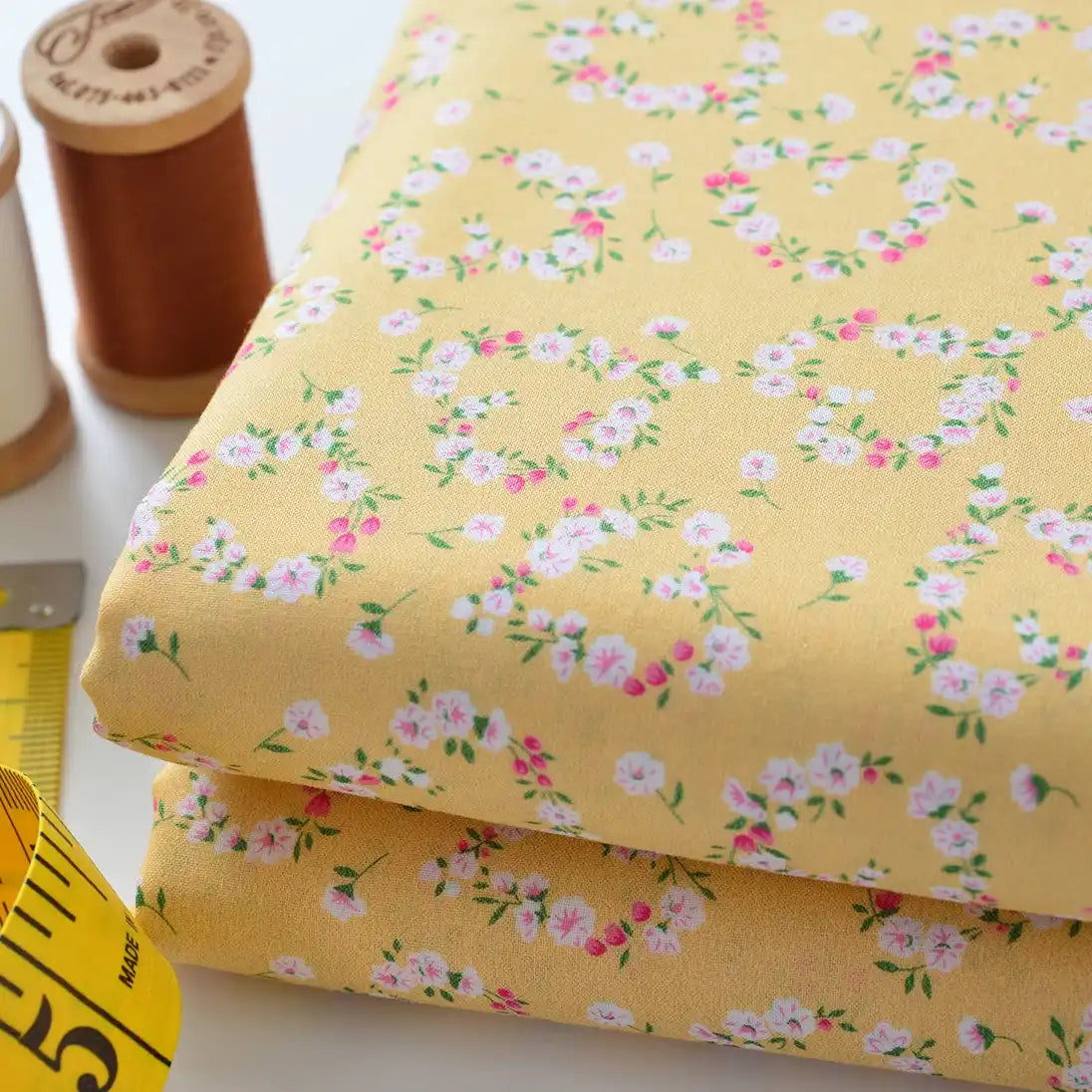 Two folded pieces of yellow Floral Lawn Fabric by Longan Craft, featuring a pink and white floral pattern, are shown with brown and white thread spools and a yellow measuring tape. This lightweight cotton lawn is ideal for breezy, breathable projects.