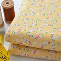 Two folded pieces of yellow Floral Lawn Fabric by Longan Craft, featuring a pink and white floral pattern, are shown with brown and white thread spools and a yellow measuring tape. This lightweight cotton lawn is ideal for breezy, breathable projects.