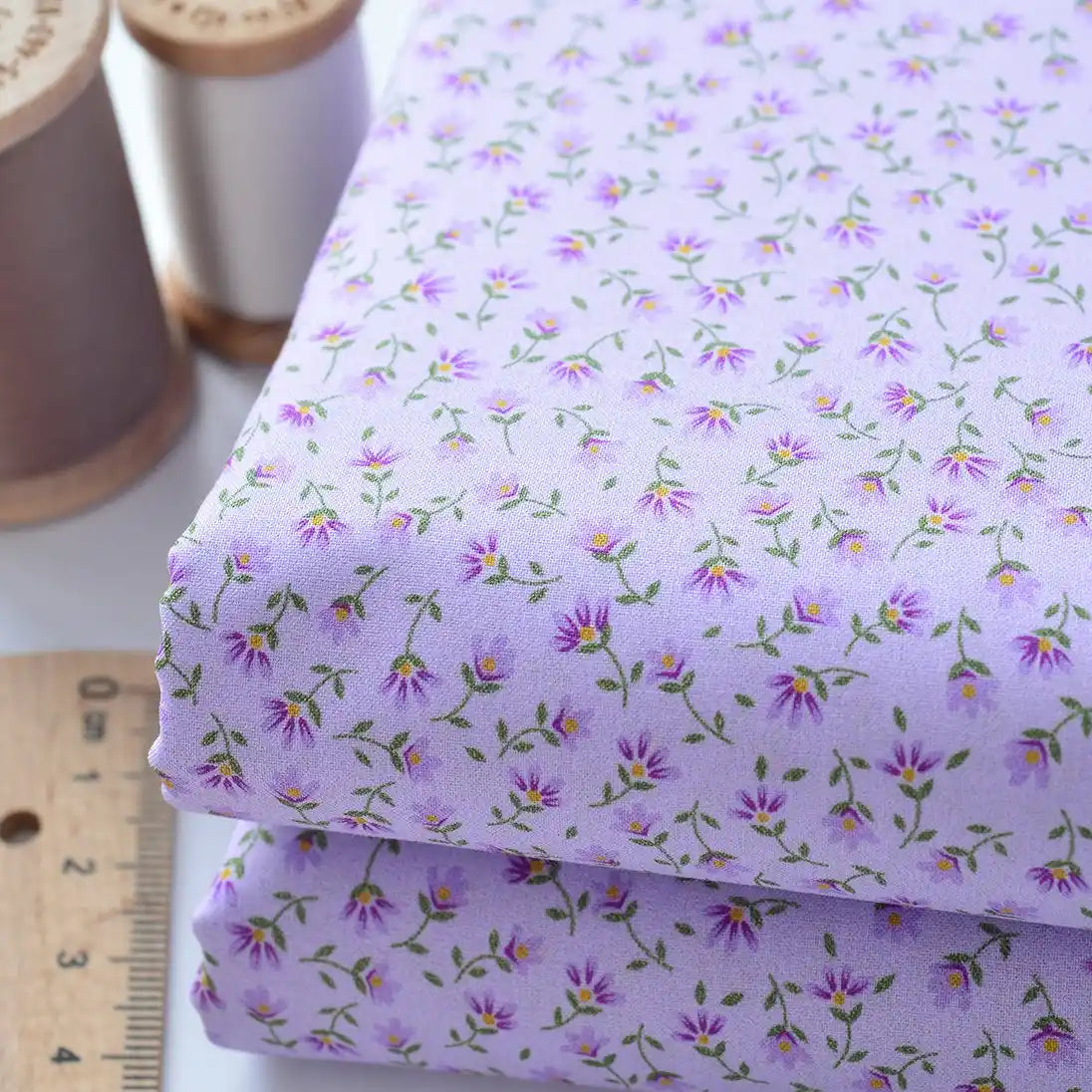 Two folded pieces of Floral Lawn Fabric By The Yard from Longan Craft, featuring a light purple base with small purple and yellow flowers, shown beside wooden spools of thread and a ruler. Lightweight cotton, perfect for breathable dress projects.