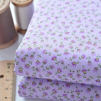 Two folded pieces of Floral Lawn Fabric By The Yard from Longan Craft, featuring a light purple base with small purple and yellow flowers, shown beside wooden spools of thread and a ruler. Lightweight cotton, perfect for breathable dress projects.