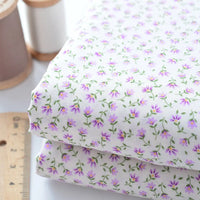 Two pieces of Longan Craft Floral Lawn Fabric By The Yard, lightweight cotton with a purple and green floral pattern, are shown alongside a ruler and spools of thread in the background.