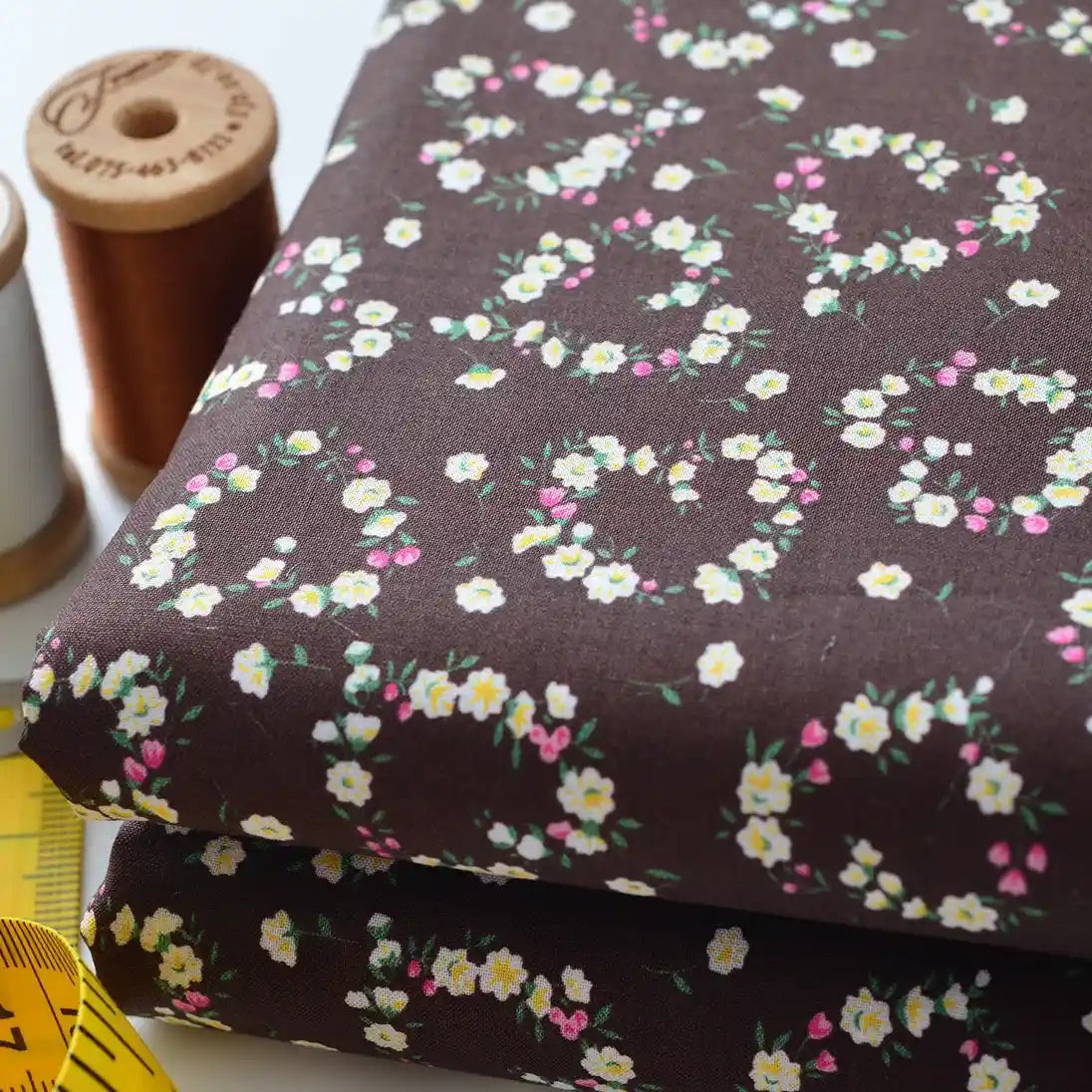 Two folded pieces of Longan Craft Floral Lawn Fabric By The Yard featuring small white, pink, and yellow floral wreaths. Shown with brown and white thread spools plus a yellow tape—ideal lightweight cotton for lining or sewing projects.