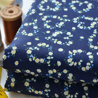 Two folded pieces of Longan Craft Floral Lawn Fabric—lightweight cotton with small white and yellow flowers—are shown beside brown spools of thread and a yellow measuring tape.