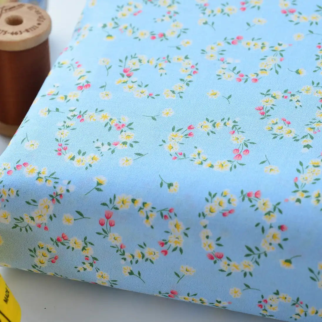 A close-up of Longan Craft’s Floral Lawn Fabric By The Yard, a lightweight, breathable cotton lawn with a yellow, white, and pink floral wreath pattern. A brown spool of thread and part of a yellow object are in the background.