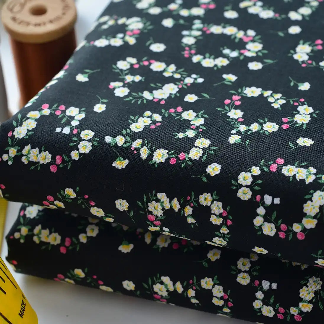 Two folded pieces of Floral Lawn Fabric By The Yard by Longan Craft, a lightweight cotton lawn with small white, yellow, and pink flowers. A spool of brown thread and a yellow measuring tape are partially visible beside the fabric.