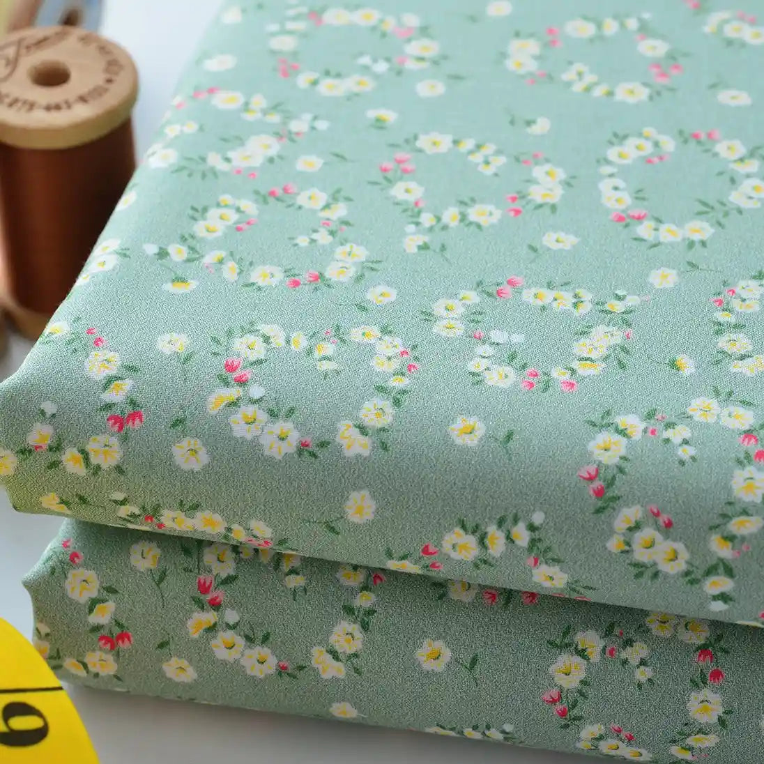 Longan Craft's Floral Lawn Fabric By The Yard features pastel green cotton with small white, yellow, and pink flowers—perfect as lightweight, breathable dress or lining fabric. Brown thread spool and yellow tape measuring shown for detail.