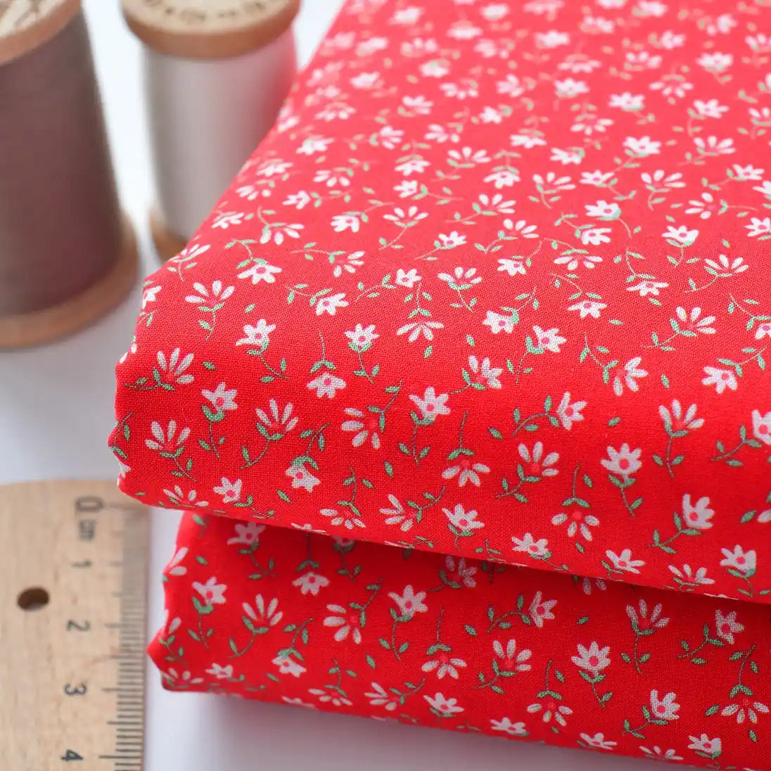 Two pieces of Longan Craft Floral Lawn Fabric By The Yard, featuring a small white floral pattern on red, are stacked beside a ruler and two spools of thread. This lightweight cotton lawn fabric is ideal for breathable dress or lining projects.